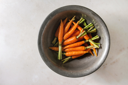 Young mini carrot in vintage metal bowl over white marble background. Flat lay, space. Cooking concept, food background.の写真素材