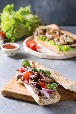 Beef baguette sandwich with tomatoes, basil, red onion served on ceramic plate with ingredients above over grey blue table.の写真素材