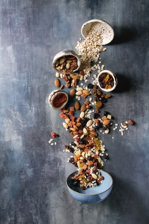 Variety of dried fruits, nuts, honey and oat flakes pours in ceramic bowl for cooking homemade healthy breakfast muesli or granola energy bars over blue texture background. Flat lay, space.の写真素材