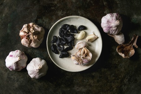 Variety of fresh organic garlic bulbs whole and peeled and cloves of black fermented garlic on ceramic plate over dark metal background. Flat lay, spaceの写真素材