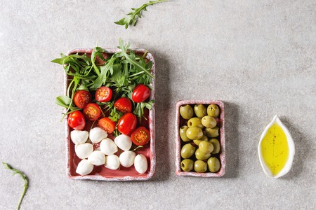 Mozzarella, cherry tomatoes, olives antipasto appetizers served in pink ceramic rectangular plates with olive oil, arugula in rowover grey texture background. Flat lay, spaceの写真素材