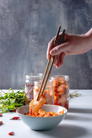 Korean traditional fermented appetizer kimchi cabbage and radish salad, fish snack served in glass jars with Vietnamese oregano and chili peppers over grey blue table. Chopsticks in mans hands.の写真素材