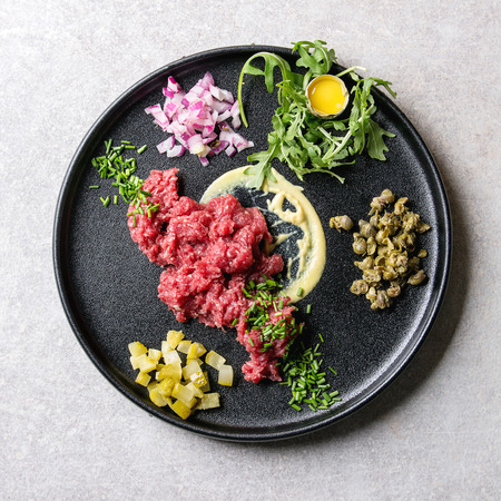 Beef tartare with quail egg in shell, cutting pickled cucumbers, capers, red onion, chives, arugula salad served in black ceramic plate over grey background. Flat lay, space. Square imageの写真素材