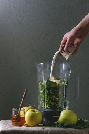 Ingredients for green spinach kale apple honey smoothie in glass blender on linen table cloth with ingredients above. Young man pouring milk. Healthy organic eating.の写真素材