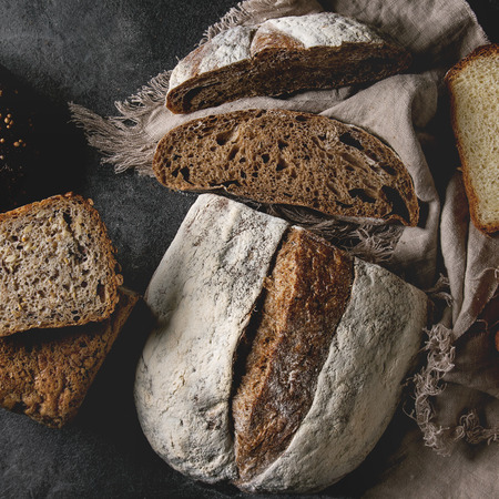 Variety of fresh baked rye, spelled, wheat craft artisan bread, whole and sliced, on cloth over black texture background. Flat lay, space. Square imageの写真素材