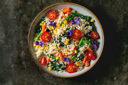 Couscous with parboiled vegetables baby carrots, green beans, sweet corn, spinach served in ceramic plate with tomatoes, sesame and edible flowers. Vegan food. Dark metal background. Flat lay, spaceの写真素材