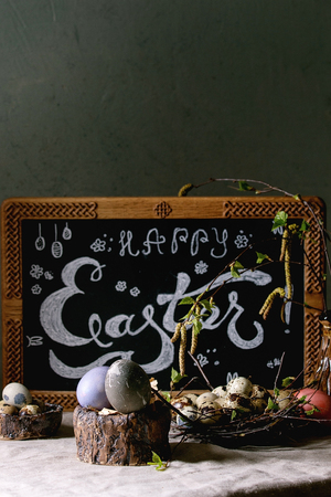 Easter holidays interior decorations. Chalkboard with Easter hand lettering, colored and quail eggs in nest, birch branches standing on grey linen table cloth with concrete wall at background.の写真素材