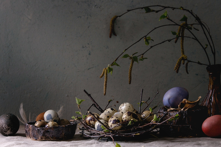 Variety of colored and quail Easter eggs in birds nest with young blooming birch branches in ceramic vase standing on grey linen table cloth with wall at background. Easter holidays decorations.の写真素材