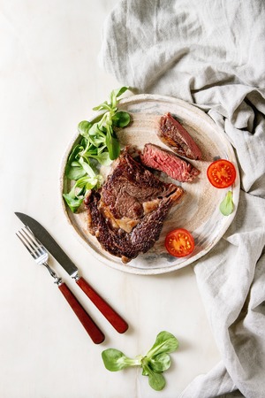 Grilled roasted sliced medium rare beef steak served in ceramic plate with green field salad, cherry tomatoes, cutlery over white marble background. Flat lay, spaceの写真素材