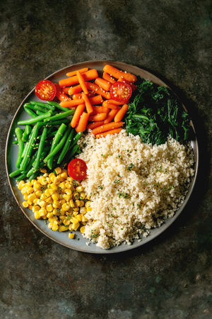 Couscous with parboiled vegetables baby carrots, green beans, sweet corn, spinach served in ceramic plate with cherry tomatoes and sesame seeds. Vegan food. Dark metal background. Flat lay, spaceの写真素材