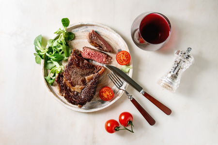 Grilled roasted sliced medium rare beef steak served in ceramic plate with green field salad, cherry tomatoes, cutlery, pepper, glass of red wine over white marble background. Flat lay, spaceの写真素材