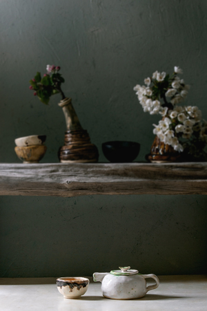 Craft handmade ceramic teapot and cup with hot tea on white marble table and spring blossom cherry branches in vase on wooden shelf.の写真素材