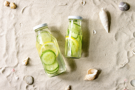 Summer drinks theme. Two glass bottles with lemon and cucumber infusion sassy water on white sand as background. Flat lay, spaceの写真素材