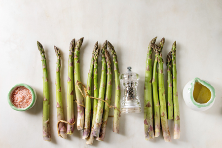 Bundle of raw organic green asparagus with cooking ingredients pink sea salt, black pepper and olive oil over white marble background. Flat lay, spaceの写真素材