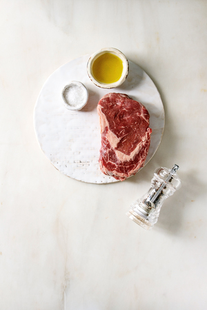Three raw uncooked beef steaks with pepper, salt and olive oil on ceramic board over white marble background. Flat lay, spaceの写真素材