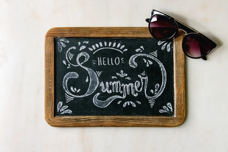 Summer theme. Handwritten decorative chalk lettering Hello summer on vintage chalkboard and sunglassesover white marble background. Flat lay, spaceの写真素材