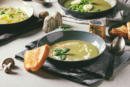 Variety of green vegetable soup asparagus, broccoli and pea, decorated by greens, vegetables, quail eggs, cream, olive oil, in ceramic bowls over grey spotted table with ingredients above.の写真素材