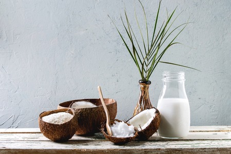 Variety of coconut products milk in glass bottle, oil and flakes in shell, fresh broken coconut on old wooden table with grey wall at background. Healthy eating, copy spaceの写真素材