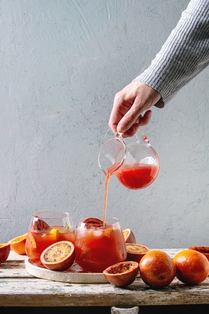 Female's hand poured from jug juice in blood orange iced cocktail in glasses, decorated by slice of oranges and fresh mint, served in ceramic tray on white wooden table with grey wall at background.の写真素材