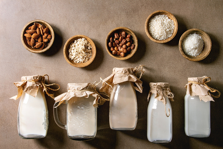 Variety of non-dairy vegan lactose free nuts and grain milk almond, hazelnut, coconut, rice, oat in glass bottles in row with ingredients above over brown texture background. Flat lay, spaceの写真素材