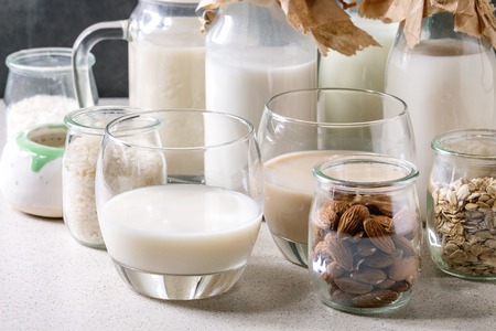 Variety of non-dairy vegan lactose free nuts and grain milk almond, hazelnut, coconut, rice, oat in glass bottles with ingredients above on white table with blue background.