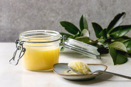 Homemade Melted ghee clarified butter in open glass jar and spoon on saucer standing on white marble table with green branch. Grey wall at background.の写真素材