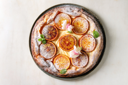 Homemade Cheesecake with sicilian blood oranges, decorated by edible flowers, mint leaves and sugar powder served in plate with cutted oranges above over white marble background. Flat lay, spaceの写真素材