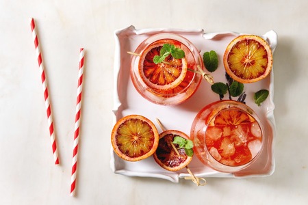 Blood orange iced cocktails in glasses, decorated by slice of oranges and fresh mint on skewers, served in ceramic tray with retro straw over white marble background. Flat lay, space.の写真素材