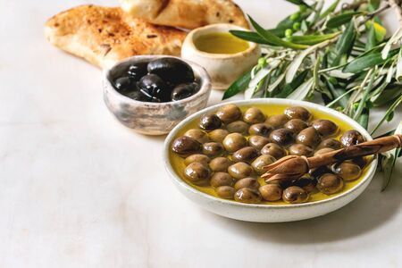 Variety of green and black whole olives in olive oil served in ceramic bowls with fresh baked ciabatta bread, pink salt and young olive wood branches on white marble table. Copy spaceの写真素材