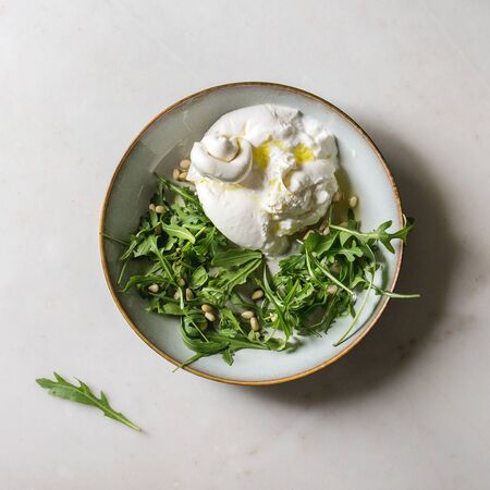 Sliced Italian burrata cheese, fresh arugula salad, pine nuts and olive oil in white ceramic plate over white marble background. Flat lay, space. Square imageの写真素材