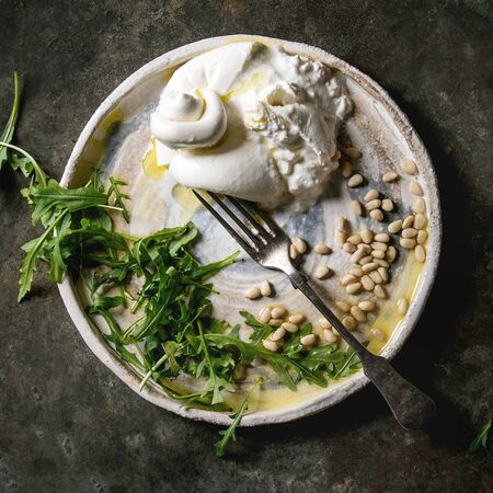 Sliced Italian burrata cheese, fresh arugula salad, pine nuts and olive oil in white ceramic plate over dark metal background. Flat lay, space. Square imageの写真素材