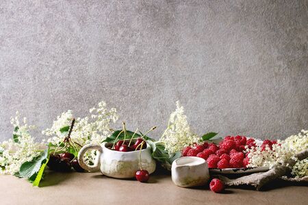 Fresh cherry and raspberry berries in ceramic mug and plate, elderflowers, jug of cream on kitchen table with gray wall at background. Copy spaceの写真素材