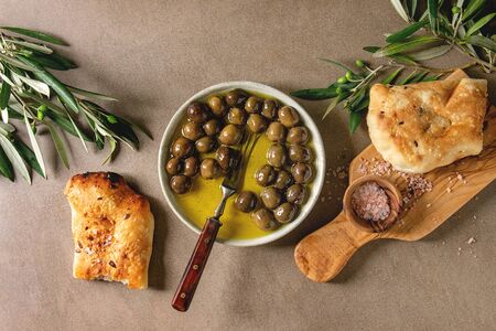 Green whole olives in olive oil served in ceramic bowl with fresh baked ciabatta bread, pink salt and young olive wood branches over brown texture background. Flat lay, spaceの写真素材