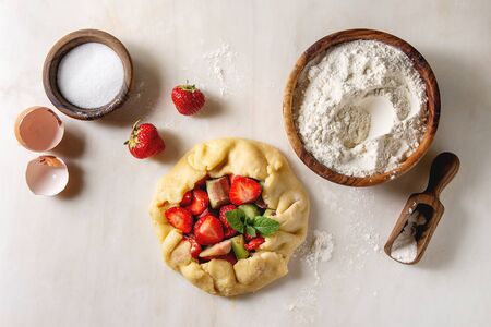 Process of baking summer berry biscuit pie. Bowl of flour, rolled shortbread dough, cutting strawberry and rhubarb, sugar over white marble background. Flat lay, spaceの写真素材