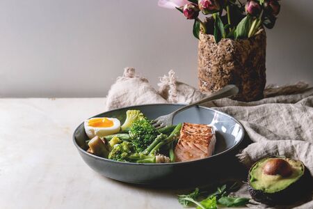 Ketogenic low carb diet dinner grilled salmon, avocado, broccoli, green bean and soft boiled egg in ceramic bowl served with olives and nuts, bouquet of peonies on grey table.の写真素材