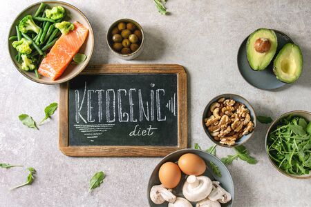 Ketogenic diet ingredients for cooking dinner. Raw salmon, avocado, broccoli, bean, olives, nuts, mushrooms, eggs in ceramic bowls with chalk board lettering. Grey texture background. Flat lay, spaceの写真素材