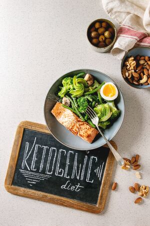 Ketogenic low carb diet dinner grilled salmon, avocado, broccoli, green bean and boiled egg in bowl served with olives, nuts and chalk board lettering over grey spotted background. Flat lay, spaceの写真素材