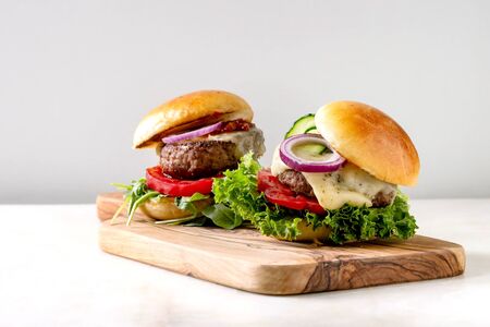 Two homemade fast food burgers classic hamburger or cheeseburger with beef, salad, cheese and tomato served on wooden cutting board on white marble kitchen table. Copy space.の素材 [FY310127280871]