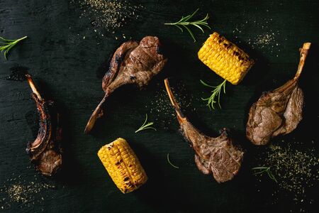 Grilled bbq rack of lamb with sweet corn cobs and rosemary over black burned wooden background. Flat...