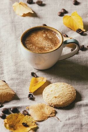 Cup of espresso coffee standing on linen table cloth with yellow autumn leaves, berries, coffee beans and shortbread cookies with dark wall at background. Fall concept. Copy spaceの写真素材