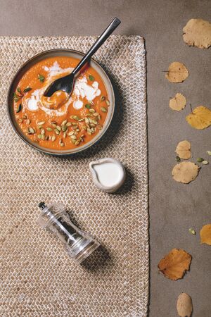 Pumpkin soup with cream and seeds in ceramic bowl, served with pepper mill and autumn leaves over brown texture background. Flat lay, spaceの写真素材
