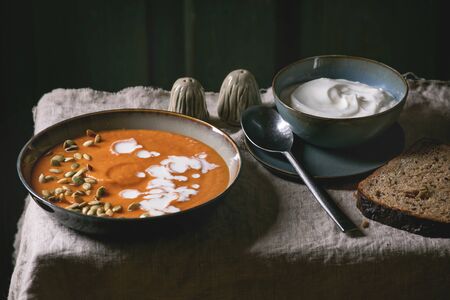 Pumpkin soup with cream and seeds in ceramic bowl, served with sour cream, rye bread, salt and pepper on grey linen tablecloth. Dark rustic styleの写真素材