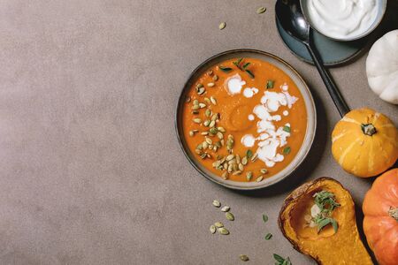 Pumpkin soup with cream and seeds in ceramic bowl, served with sour cream, baked and uncooked pumpkins over brown texture background. Flat lay, spaceの写真素材