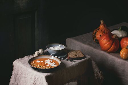 Pumpkin soup with cream and seeds in ceramic bowl, served with sour cream, rye bread, uncooked pumpkins on grey linen tablecloth. Dark rustic styleの写真素材