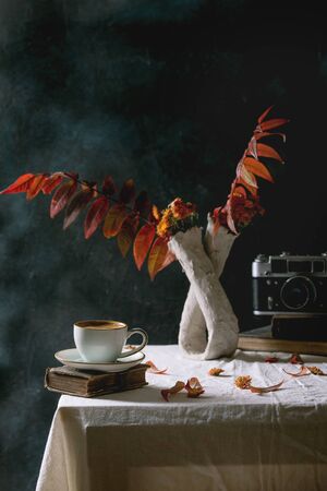 Cup of black coffee espresso standing on white table cloth in dark room with autumn leaves and flowers in clay vase, old books, vintage camera.の写真素材