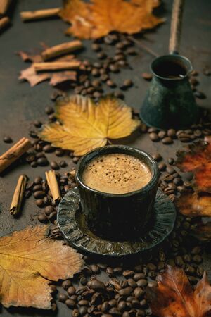 Black coffee espresso with foam in black ceramic cup, with saucer, cezve coffee pot, autumn leaves, spices and roasted beans above over brown texture background.の写真素材