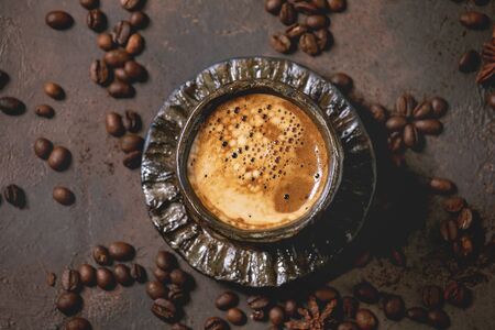 Black coffee espresso with foam in black ceramic cup, with saucer and roasted beans above over brown texture background. Flat lay, spaceの写真素材