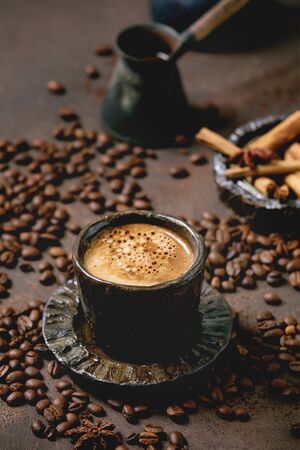 Black coffee espresso with foam in black ceramic cup, with saucer, cezve coffee pot, spices and roasted beans above over brown texture background.の写真素材