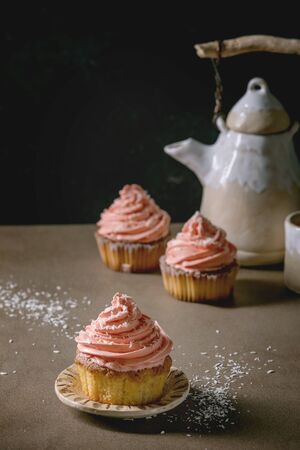 Homemade cupcake with pink buttercream and coconut flakes served with ceramic teapot, cup of tea on dark table.の写真素材