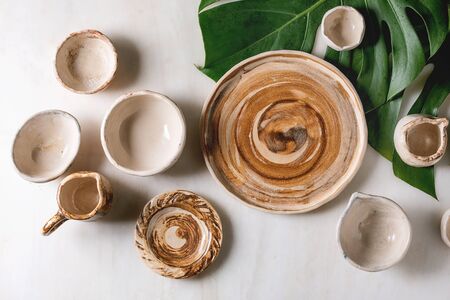 Variety of empty different ceramic dishes, bowls, jugs and plates on exotic monstera leaves over white marble background. Flat lay, spaceの写真素材